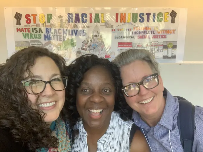 Photo of Mollie, Fernell, and Jen. They're smiling while looking at the camera for a selfie. Behind them is a poster with a header which reads stop racial injustice.