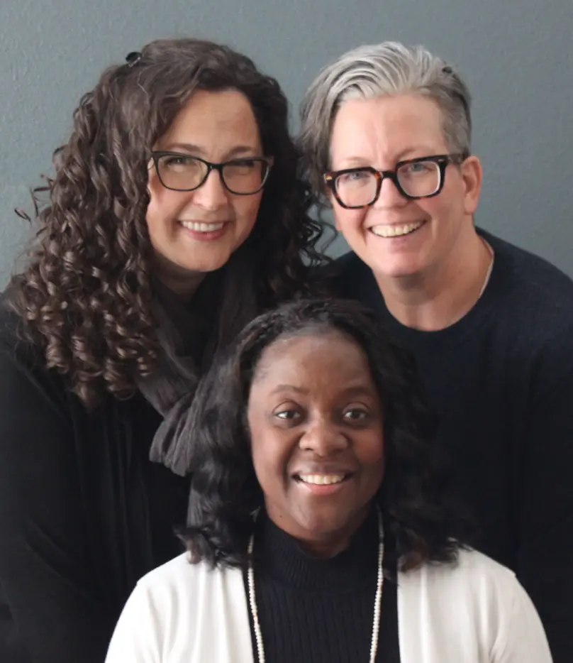 Photo of Mollie, Fernell, and Jen. Mollie and Jen are standing behind a seated Fernell. They are smiling while looking directly at the camera.